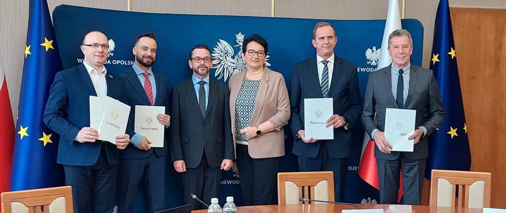 Na zdjęciu znajduje się Wojewoda Monika Jurek, Dyrektor Wydziału Infrastruktury i Nieruchomości Marek Świetlik oraz przedstawiciele gmin Głuchołazy, Zawadzkie, Jemielnica oraz Namysłów. Za nimi widać granatową ściankę z napisami "Wojewoda Opolski". Po lewej i prawej stronie widać flagi Polski oraz Unii Europejskiej. Przed nimi widać stół, na którym leży między innymi laptop, dokumenty czy butelki wody. 