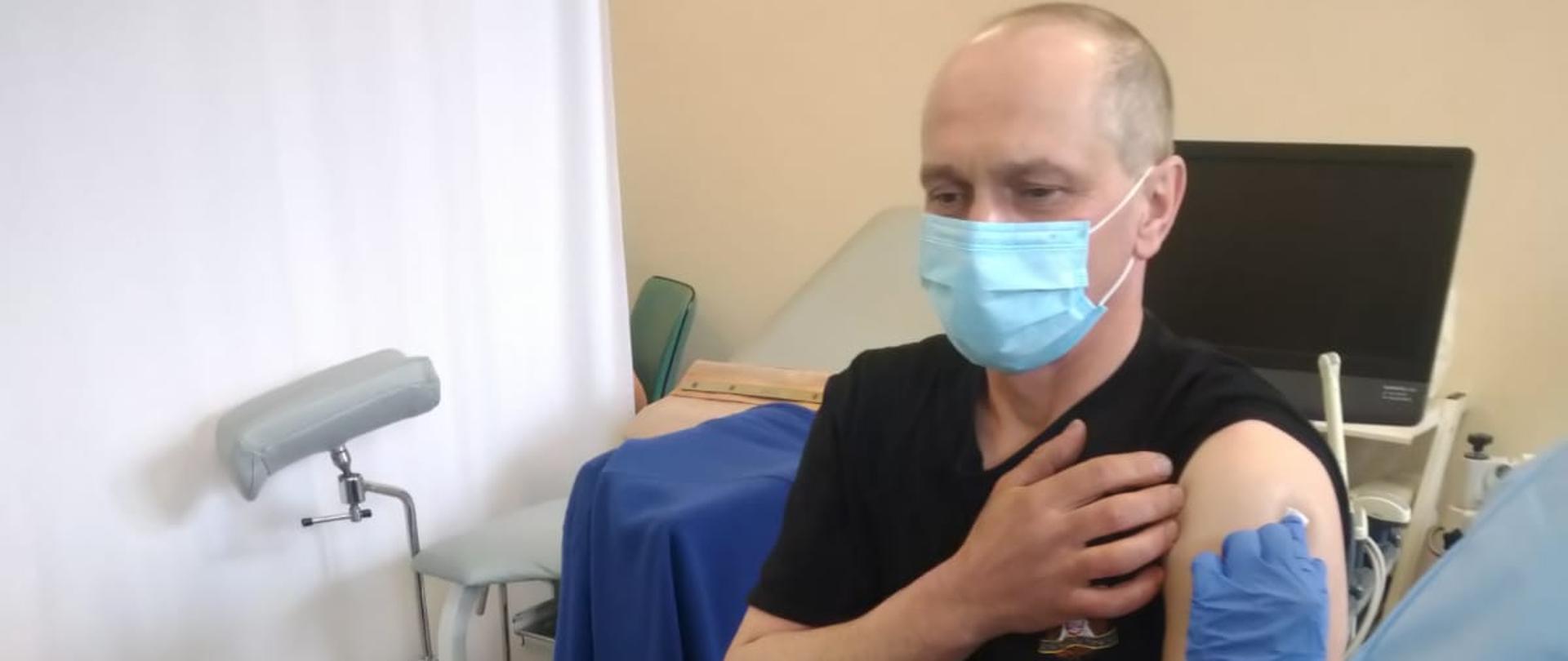 Na zdjęciu strażak w gabinecie medycznym, siedzi na ławce, ubrany w czarne spodnie koszarowe oraz czarną koszulkę z maseczką na twarzy. W lewy ramię dostaje szczepionkę.
