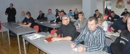Narada operacyjno-szkoleniowa. W świetlicy komendy odbywa się narada z udziałem pracowników urzędów, komendantów gminnych i przedstawicieli OSP. Uczestnicy oglądają na ekranach przygotowaną prezentację multimedialną. Naradzie przewodniczy zastępca komendanta powiatowego PSP. Z prawej strony widoczne logo PSP.