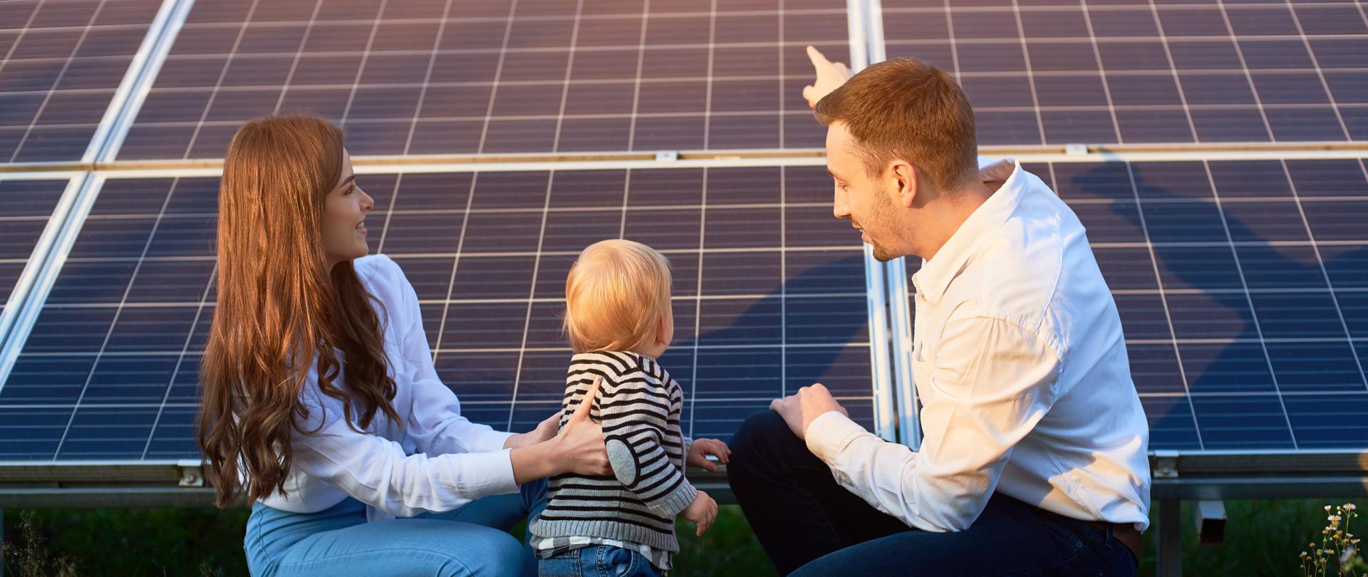 Father shows his family the solar panels on the plot near the house during a warm day. A young girl with a child and a man in the sun rays look at the solar panels.