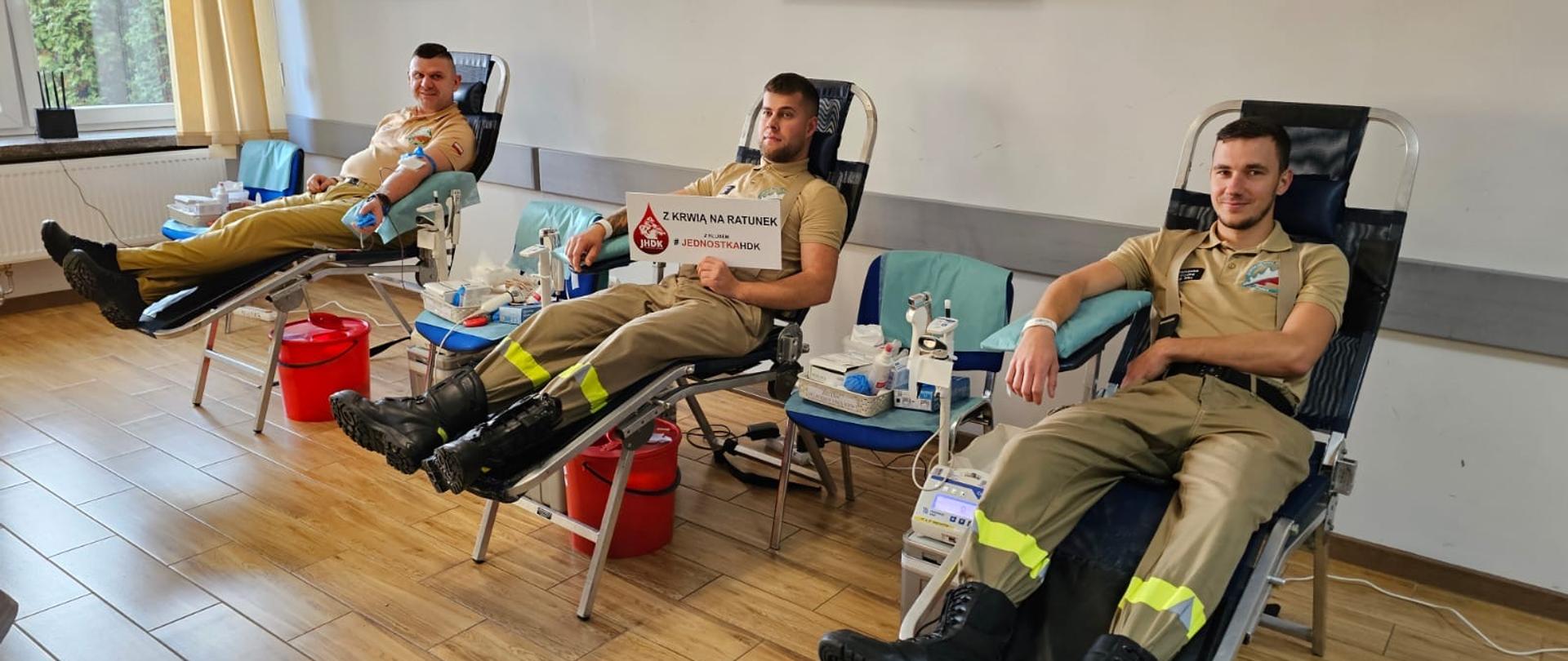 Trzech strażaków w mundurach oddaje krew, leżąc na specjalnych fotelach w sali. Każdy z nich ma podłączony zestaw do pobierania krwi. Jeden z mężczyzn trzyma tabliczkę z napisem „Z KRWIĄ NA RATUNEK – #JEDNOSTKAHDK”. W tle widać okna i elementy wyposażenia sali.