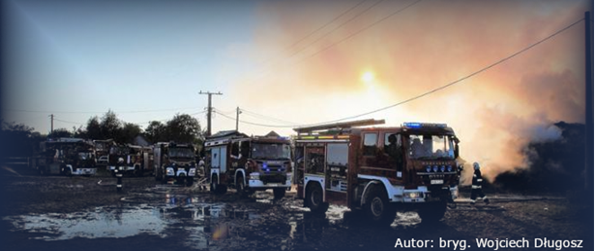 Na zdjęciu widzimy sześć samochodów pożarniczych ciężarowych z których są rozwinięte linie gaśnicze, widać chodzących strażaków w pełnym uzbrojeniu. W tle widać dymiące się sterty gaszonego siana.