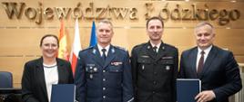 Przed napisem „Województwa Łódzkiego” cztery osoby pozują do wspólnego zdjęcia, trzymając w dłoniach podpisane egzemplarze dokumentów. W centrum stoją przedstawiciele służb mundurowych – PSP i Policji, po bokach reprezentanci Zarządu Województwa Łódzkiego. W tle flagi oraz godło państwowe.