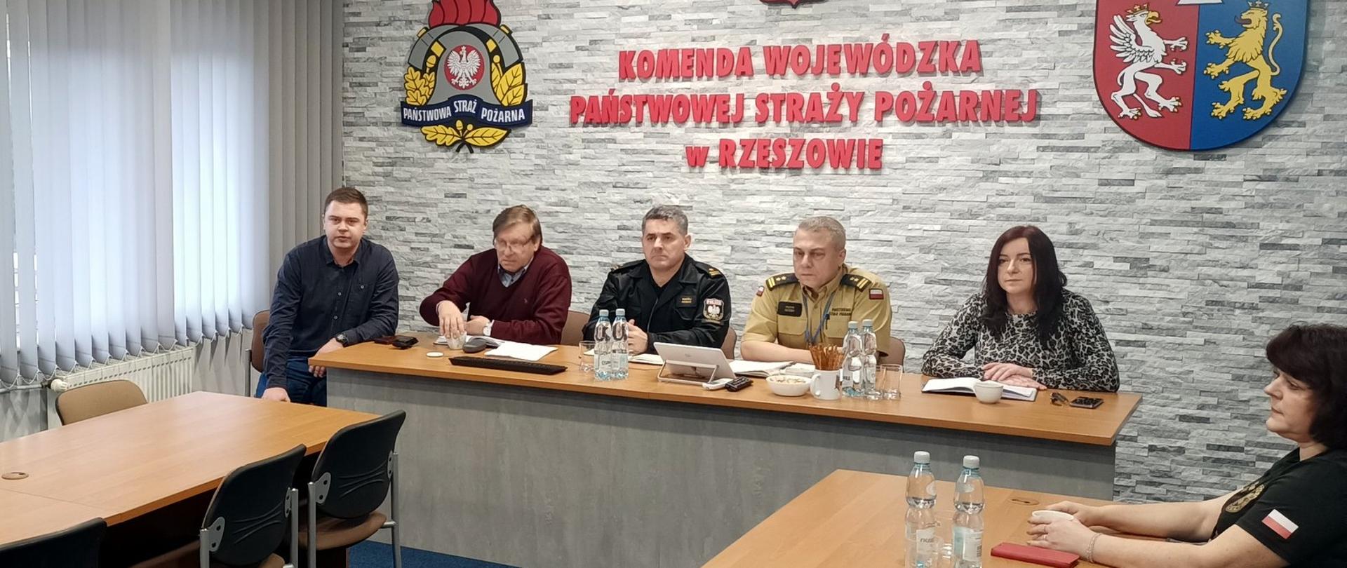 Zdjęcie zrobione w sali szkoleniowej. Na zdjęciu widać 6 osób siedzących przy stołach konferencyjnych. Wśród osób uczestniczących w szkoleniu jest dwóch funkcjonariuszy Państwowej Straży Pożarnej w mundurach. Na ścianie, za osobami siedzącymi przy stole widać napis Komenda Wojewódzka Państwowej Straży Pożarnej w Rzeszowie. Po lewej stronie napisu umieszczone jest logo Państwowej Straży Pożarnej. Po prawej stronie napisu widać część herbu województwa podkarpackiego.
