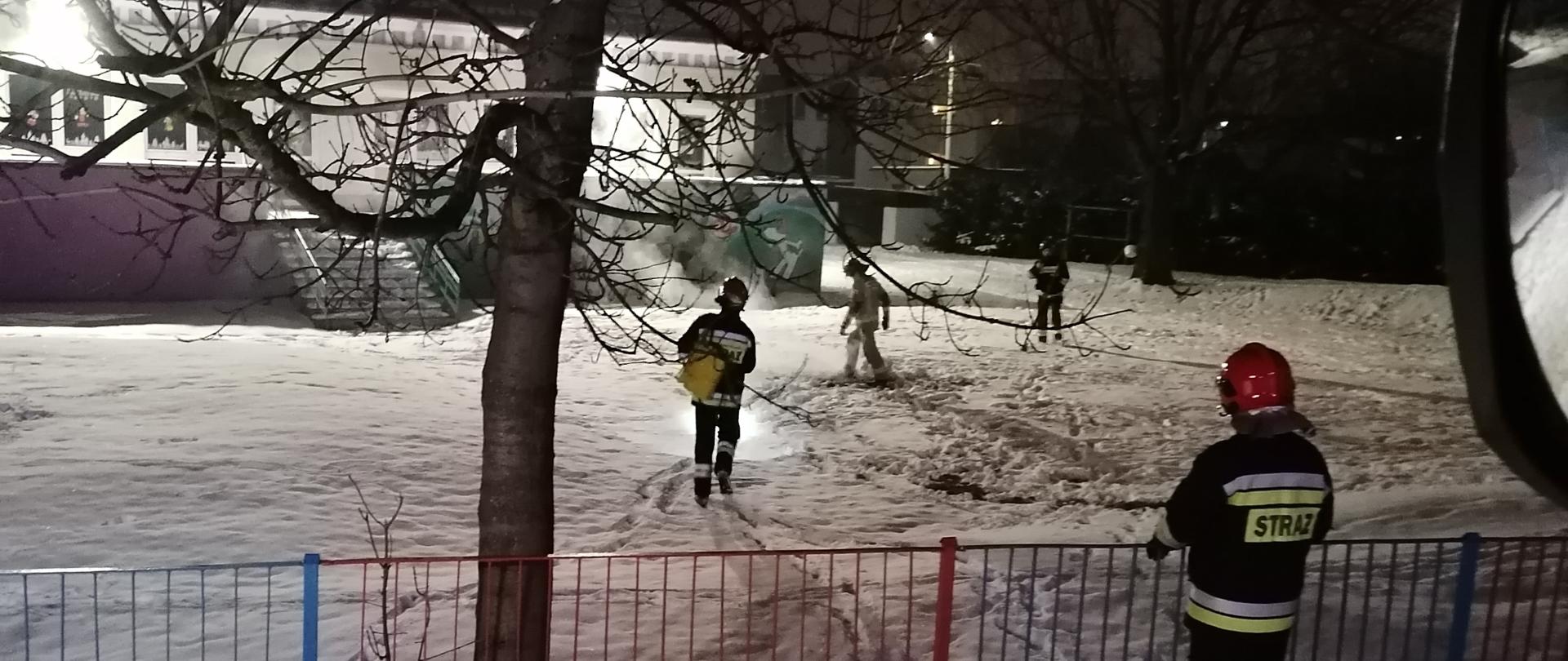 Zdjęcie przedstawia strażaków, którzy gaszą ognisko na terenie przedszkola. W tle budynek przedszkola oraz biały dym po ugaszeniu ogniska