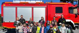 Wspólne zdjęcie grupy przedszkolaków wraz z przedszkolankami i strażakami państwowej straży pożarnej w ubraniach koszarowych na tle samochodu ratowniczo-gaśniczego.
