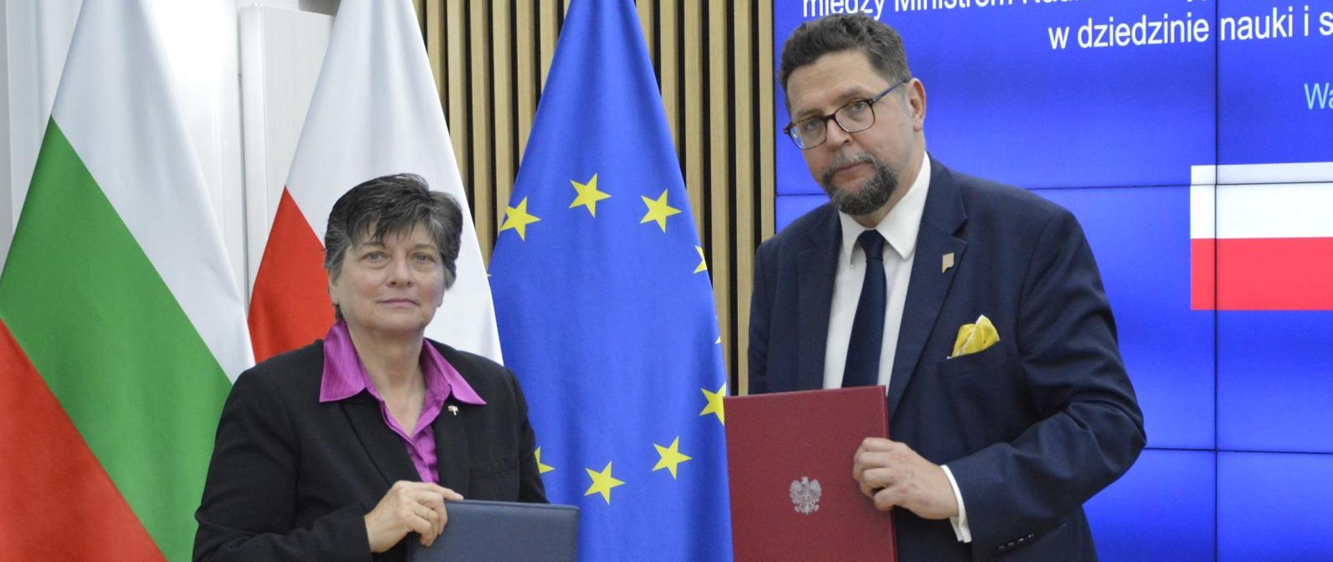 Pod ścianą przy której stoją flagi Polski, Bułgarii i UE stoi wiceminister Szeptycki ikobieta w czarnej marynarce, pokazują trzymane w rękach teczki z dokumentami.