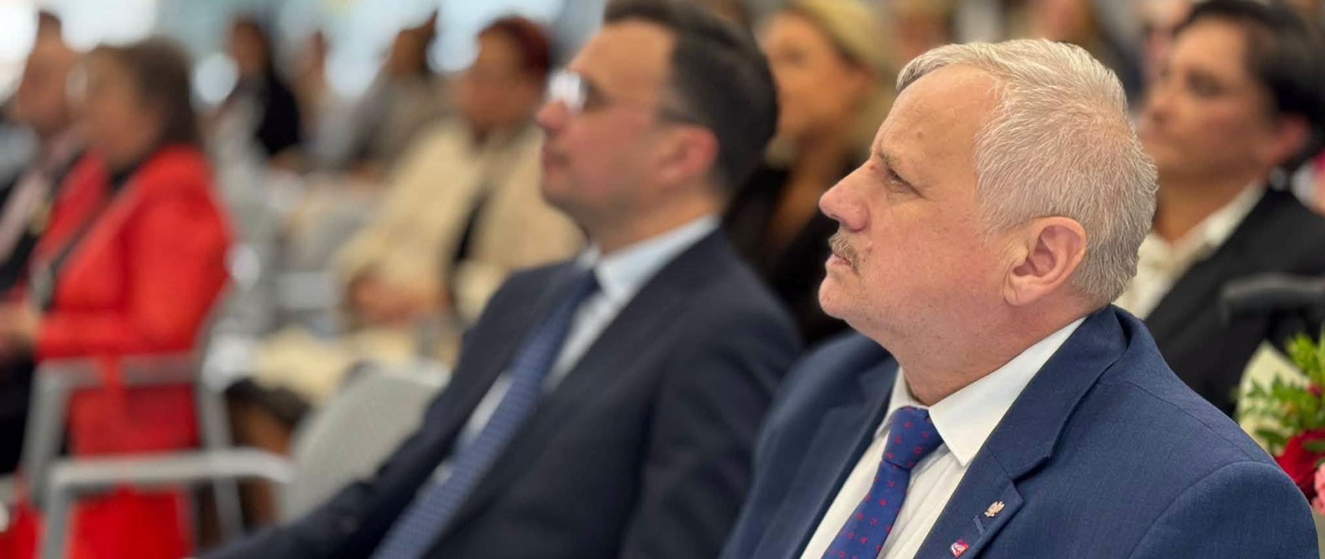 Na pierwszym planie wicewojewoda w granatowym garniturze. W tle widoczni, w lekkim rozmyciu, uczestnicy wydarzenia siedzący na widowni.