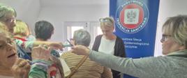 grupa osób stoi przy stoliku z ulotkami a przed nimi stoi pracownica promocji zdrowia i rozdaje ulotki w tle baner w kolorze niebieskim na którym nadrukowane jest logo inspekcji sanitarnej oraz napis Powiatowa Stacja Sanitarno-Epidemiologiczna w Szczytnie