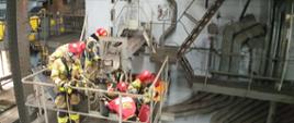 Zdjęcie wewnątrz elektrociepłowni. Oświetlenie naturalne. Widać piece oraz podesty techniczne, stalowe. Na zdjęciach ratownicy w kombinezonach wysokościowych. Widać sprzęt ratownictwa wysokościowego.