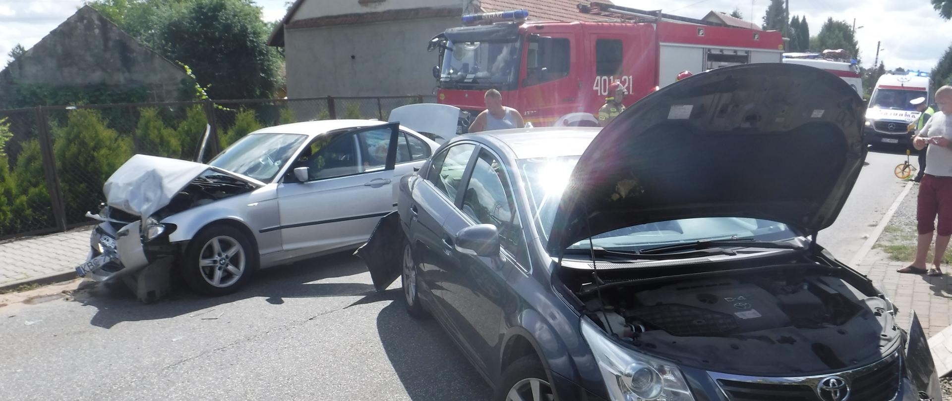 Zdjęcie przedstawia wypadek dwóch samochodów osobowych marki Toyota i BMW. Pojazdy znajdują się na drodze asfaltowej. Samochód Toyota ma otwartą klapę przednią, uszkodzoną lewą lampę przednią. na tyle pojazdu uszkodzony jest zderzak tylni. Obok pojazdu Toyota stoi mężczyzna ubrany w czerwone spodenki i koszulkę białą, a za nim widać funkcjonariusza Policji i pojazd Zespołu Ratownictwa Medycznego samochodem. Obok pojazdu Toyota stoi samochód marki BMW ma rozbity przód i pogiętą maskę przednią oraz otwarte drzwi przednie i tylną klapę bagażnika. Za samochodem BMW stoi pojazd pożarniczy w kolorze czerwonym z włączonymi niebieskimi światłami błyskowymi i odsuniętą roletą środkową zabudowy pojazdu. Za pojazdem BMW stoi mężczyzna a obok niego strażak w piaskowym ubraniu specjalnym i czerwonym hełmie.