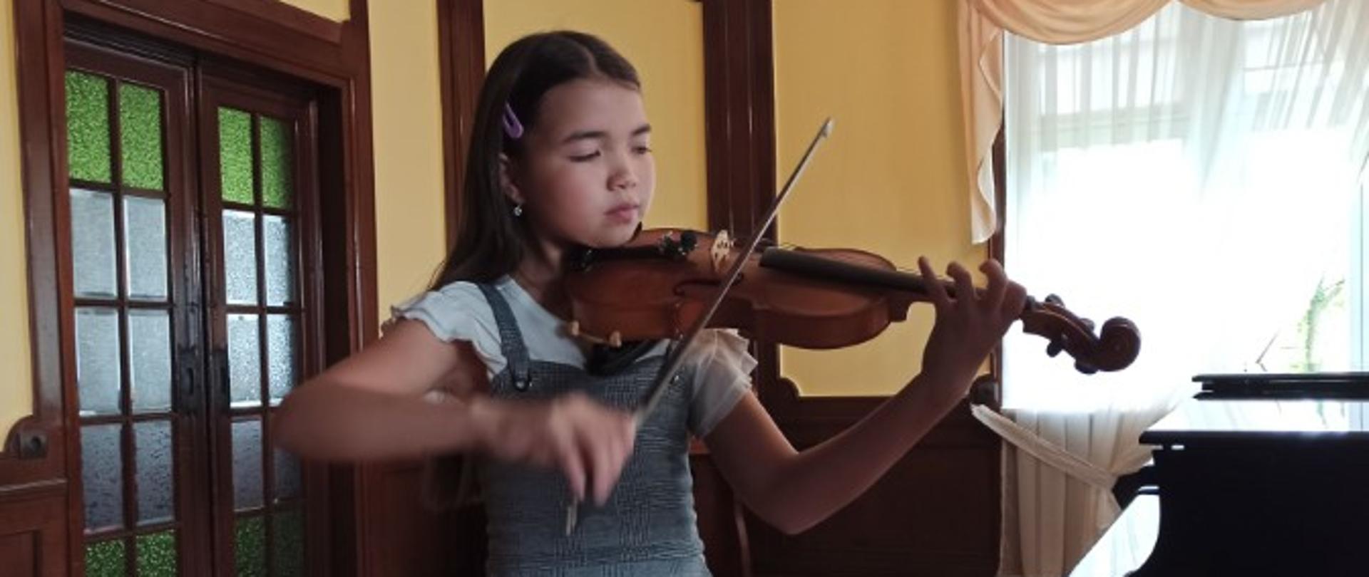 Zdjęcie kolorowe. Ciemnowłosa dziewczynka w biało- szarej sukience gra na skrzypcach. W tle fortepian.