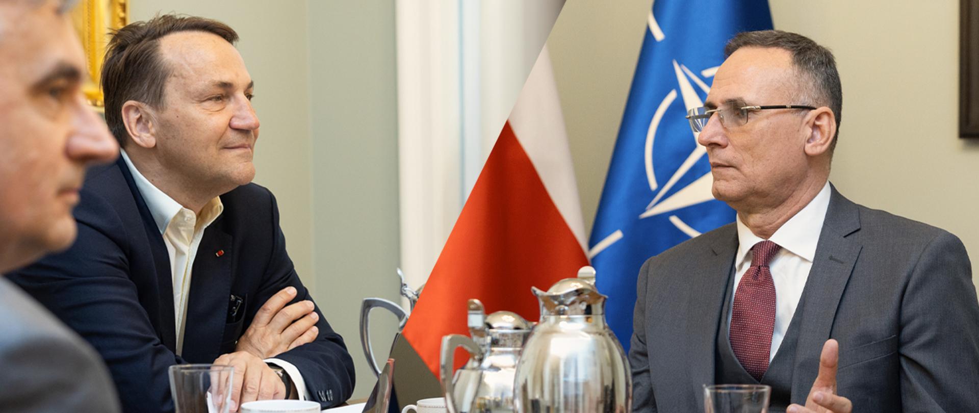 Na zdjęciu jest dwóch mężczyzn w garniturach. Siedzą przy stole, na którym są szklanki i srebrne dzbanki. Za nimi są flagi: Polski NATO