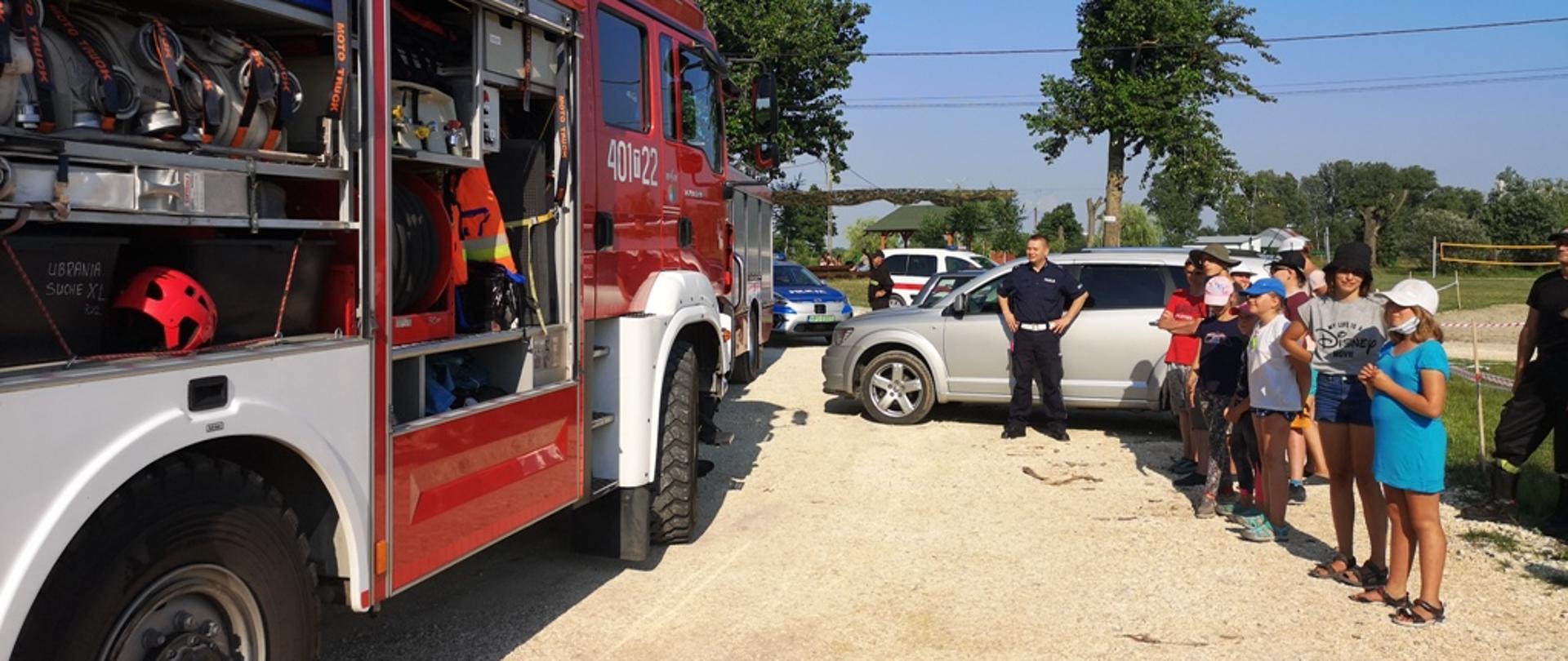 Na zdjęciu pojazd pożarniczy przed którym stoją harcerze, w tle policjant oraz strażak. 