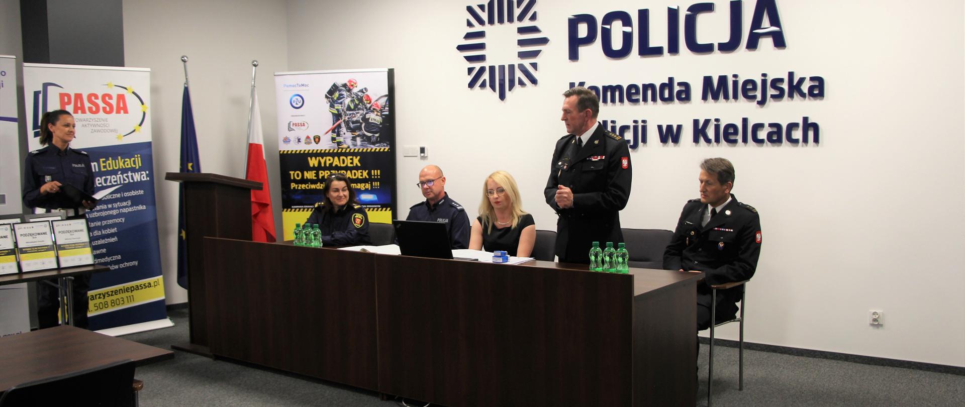 Na zdjęciu: za stołem siedzą organizatorzy projektu. St. bryg. Sławomir Karwat zabiera głos. Nad nimi znak graficzny i napis: Policja Komenda Miejska Policji w Kielcach . Po lewej stronie prelegentka za pulpitem.