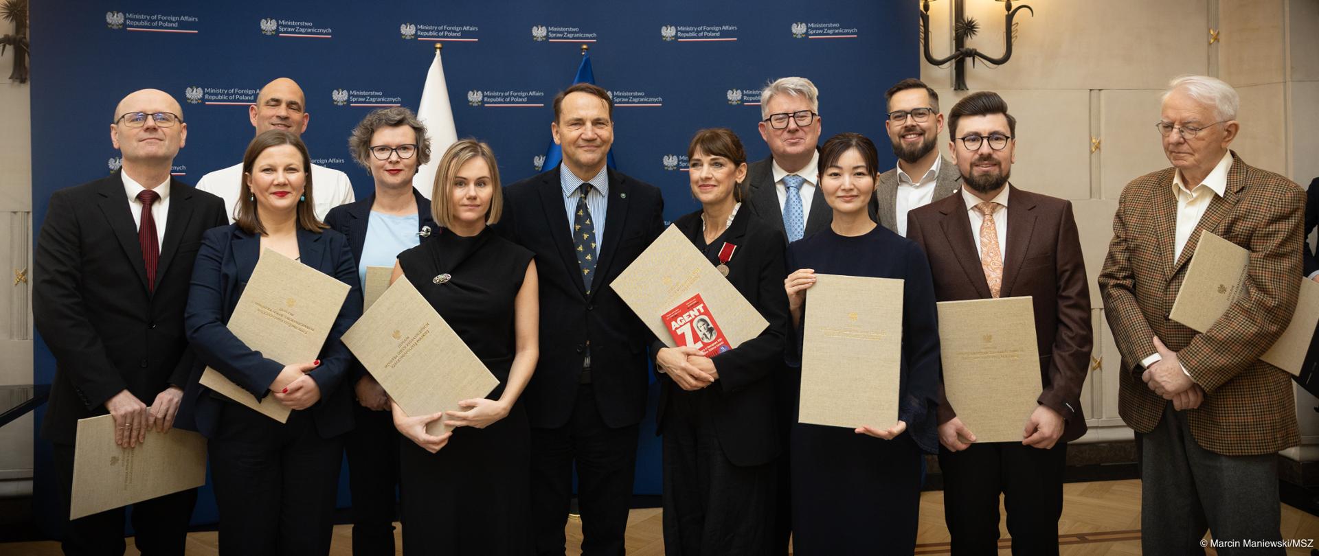 A group of people stands with Deputy PM, FM Radosław Sikorski in front of a blue government backdrop, each holding a certificate, gathered for a Best History Book Prize ceremony. 