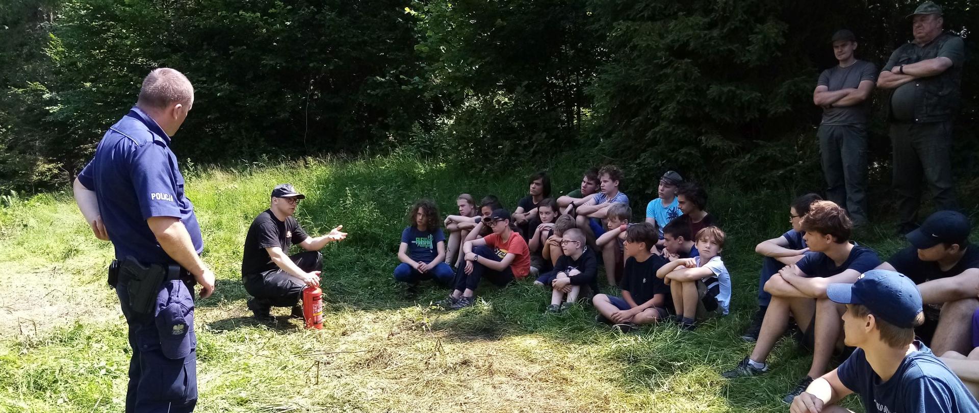 strażak psp prowadzi prelekcję dla harcerzy na polanie leśnej w obecności policjanta