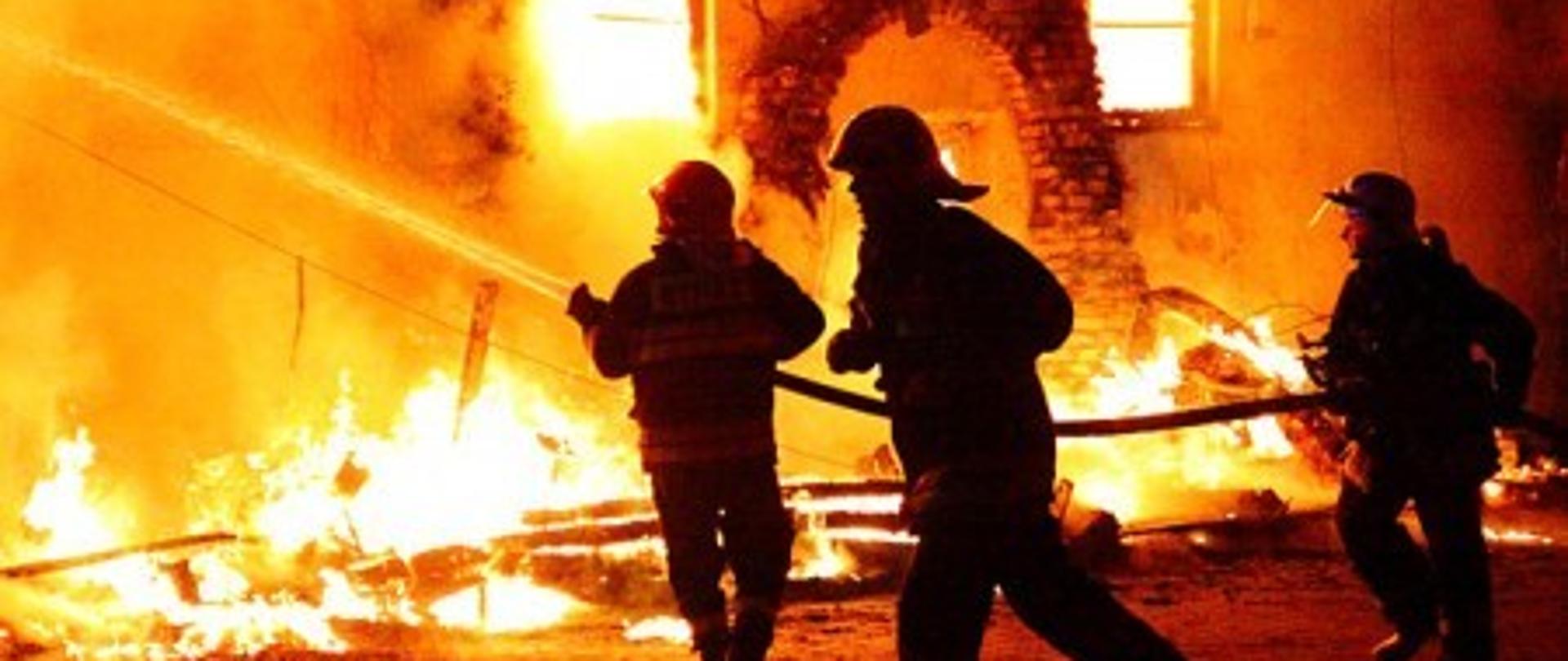 Na zdjęciu znajduje się trzech strażaków gaszących płonący budynek.