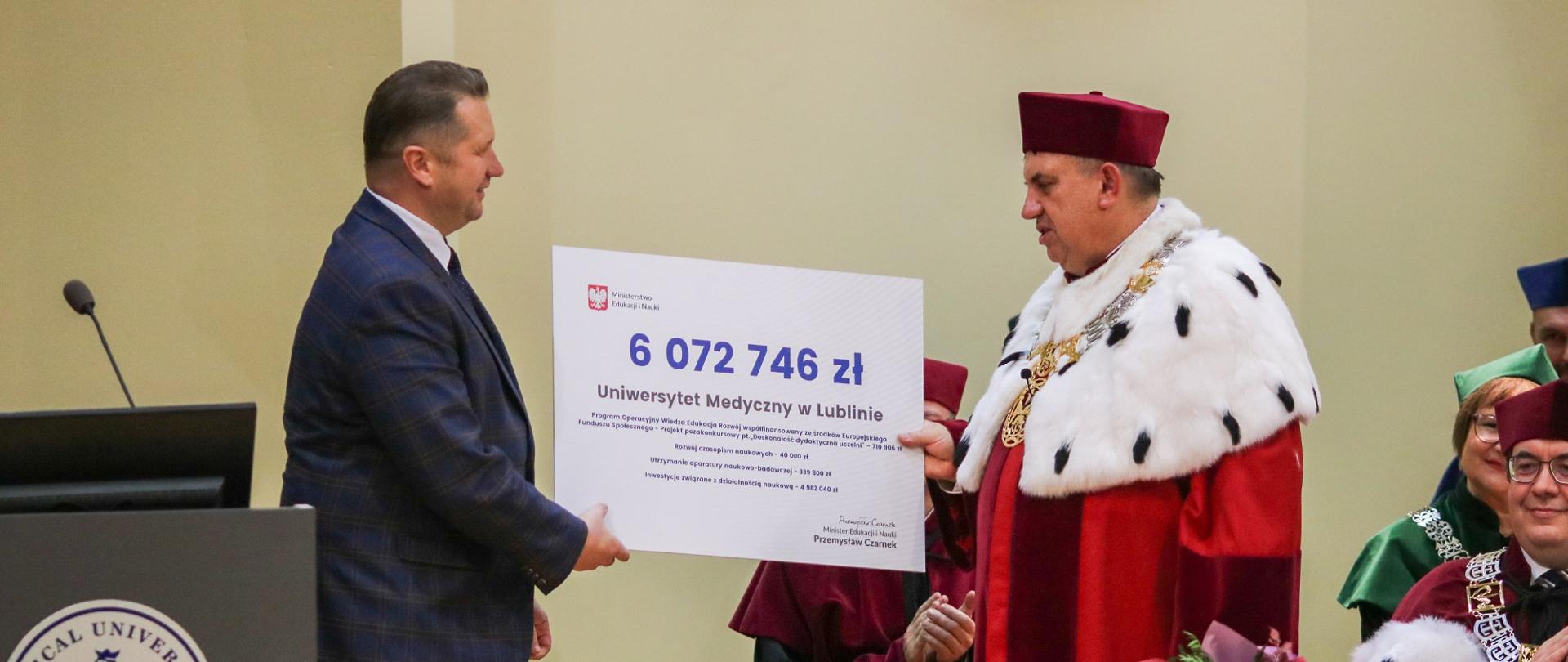 Minister Czarnek podaje mężczyźnie w ceremonialnym czerwonym stroju z gronostajowym kołnierzem wielki symboliczny czek z napisem 6 072 746 zł.