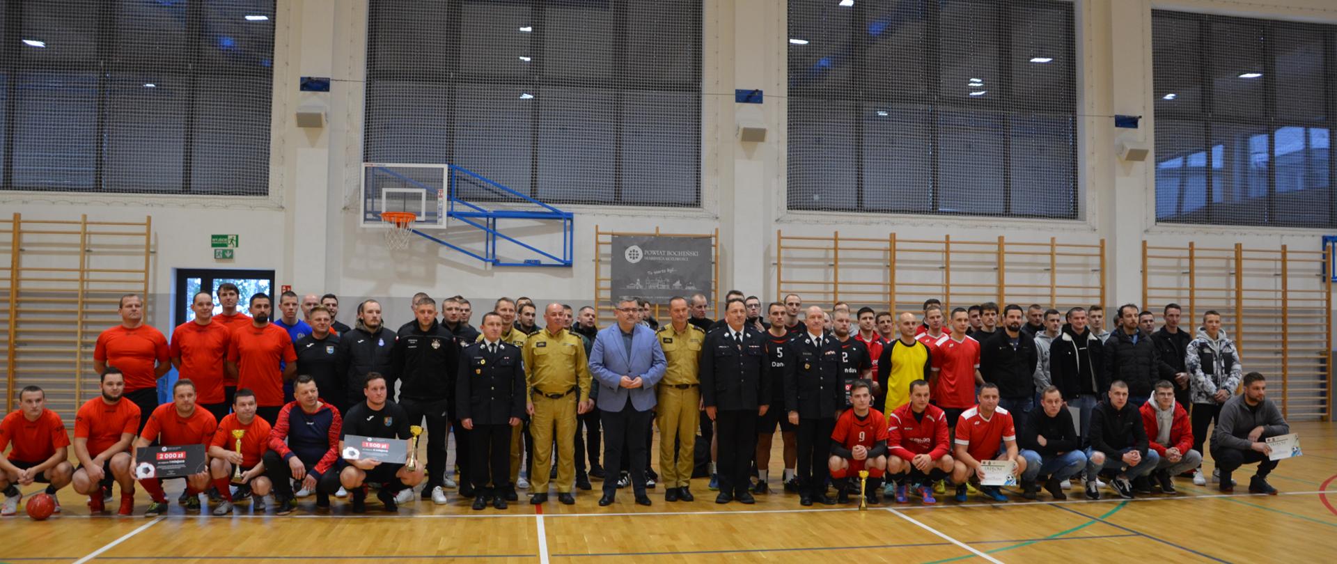 Zdjęcie przedstawia kilkudziesięciu strażaków ubranych w ubrania sportowe krótkie spodenki i koszulki z krótkim rękawem, umundurowanie służbowe koloru żółtego, umundurowanie wyjściowe koloru granatowego. W tle ściana na której zamontowane są drabinki drewniane, okna