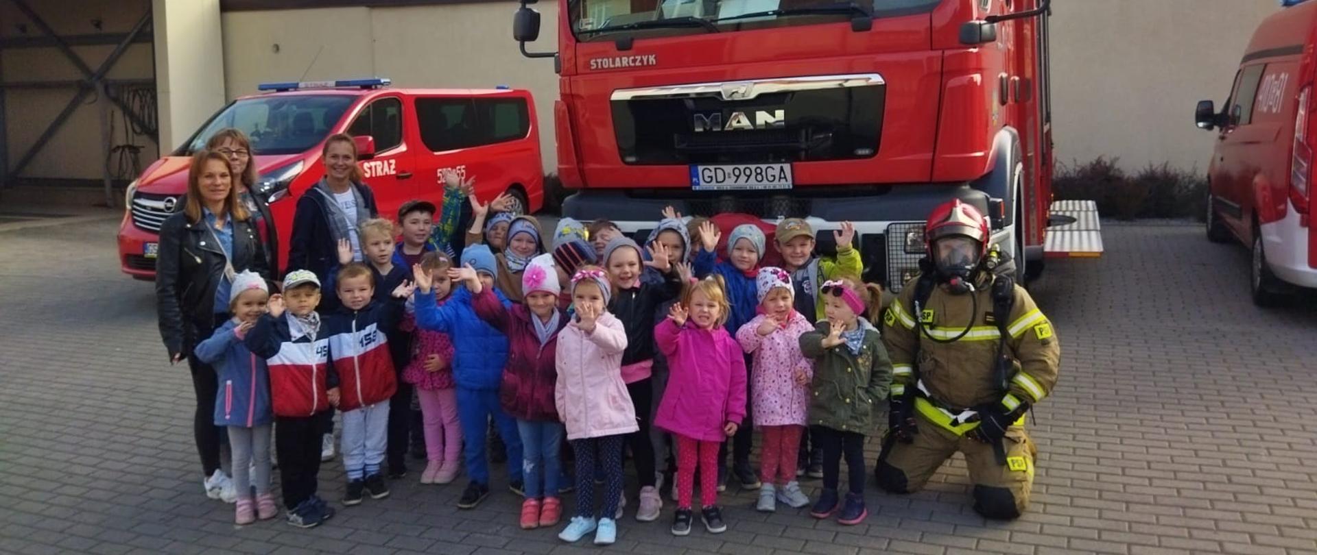 Zdjęcie przedstawia cała grupę dzieci wraz z wychowawcami na tle samochodu pożarniczego, Strażak ubrany w sprzęt ochrony dróg oddechowych i hełm.
