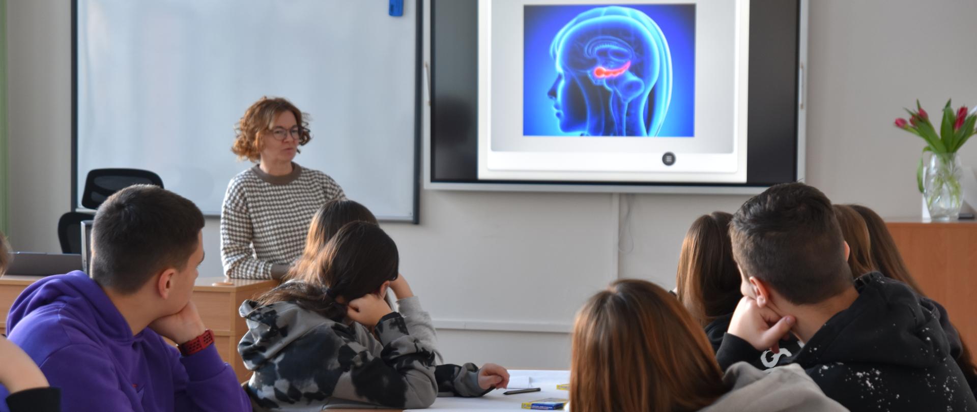Nauczycielka prowadzi lekcję w klasie szkolnej, stojąc przed ekranem z prezentacją, podczas gdy uczniowie siedzą w ławkach i uważnie słuchają.