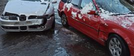Zdjęcie przedstawia dwa pojazdy uczestniczące w zdarzeniu widoczne od przodu. Z lewej strony srebrny Seat z uszkodzoną maską przednią i atrapą chłodnicy. Z prawej czerwone BMW z widocznymi wgnieceniami nadkola i drzwi przednich pasażera. Pojazdy obsypane śniegiem. Za pojazdem BMW widoczny od pasa w górę strażak w ubraniu specjalnym i czerwonym hełmem. W tle zdjęcia widoczne domy mieszkalne po obu stronach drogi