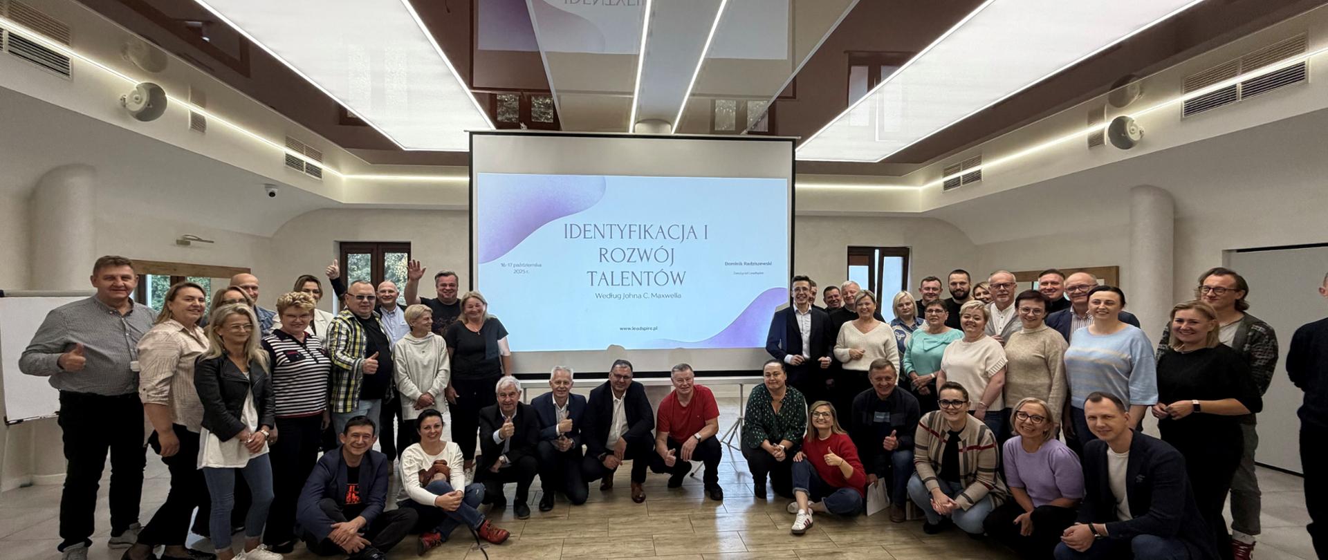 Duża grupa osób – kierowników mazowieckich placówek KRUS – pozuje w eleganckiej sali do zdjęcia grupowego. W centralnej części kadru duża plansza, na której rzutnik wyświetla slajd tytułowy prezentacji z napisem „Identyfikacja i rozwój talentów według Johna C. Maxwella”.