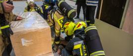 Strażacy w ubraniach specjalnych udzielają pierwszej pomocy osobie poszkodowanej podczas ćwiczeń w budynku produkcyjnym. W tle strażak w kamizelce ostrzegawczej oceniający działania. W tle widać użyte opatrunki.