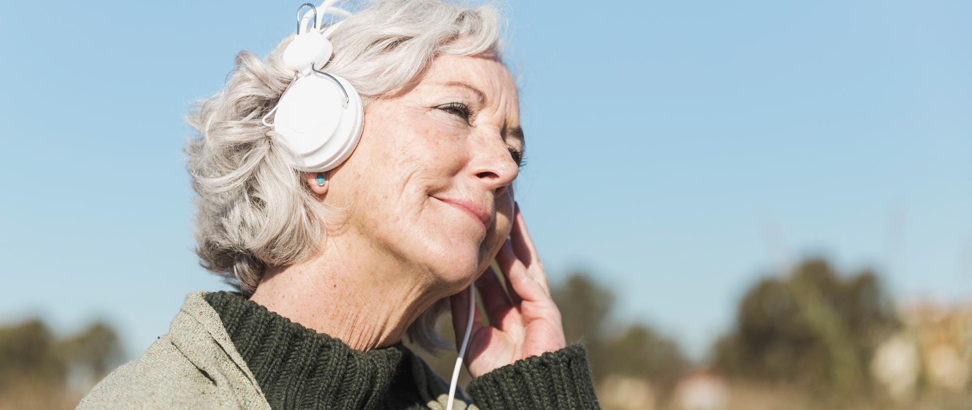 starsza kobieta słucha muzyki przez słuchawki siedząc na łonie natury