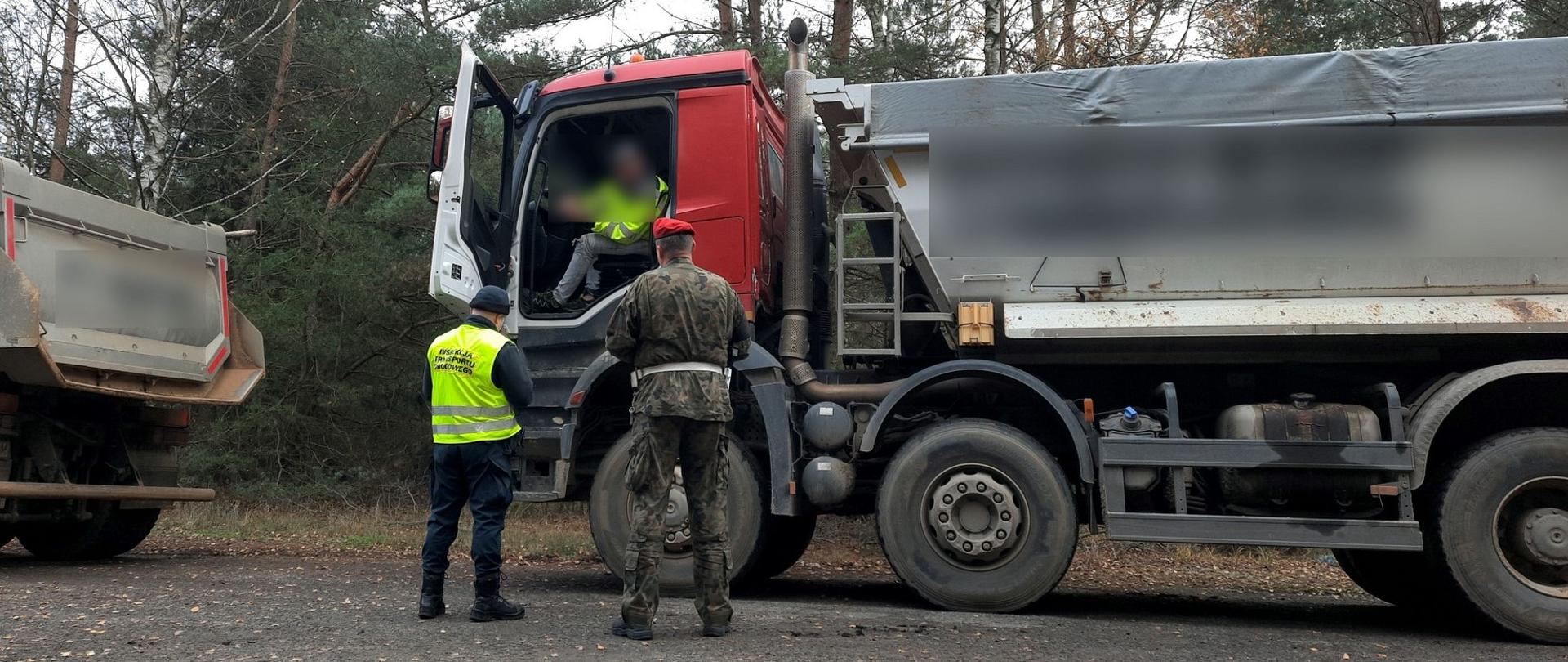 Umundurowany funkcjonariusz z wielkopolskiej Inspekcji Transportu Drogowego i Żandarmerii Wojskowej stoją obok kabiny kontrolowanej wywrotki. Inspektor weryfikuje dokumenty okazane przez kierowcę ciężarówki.