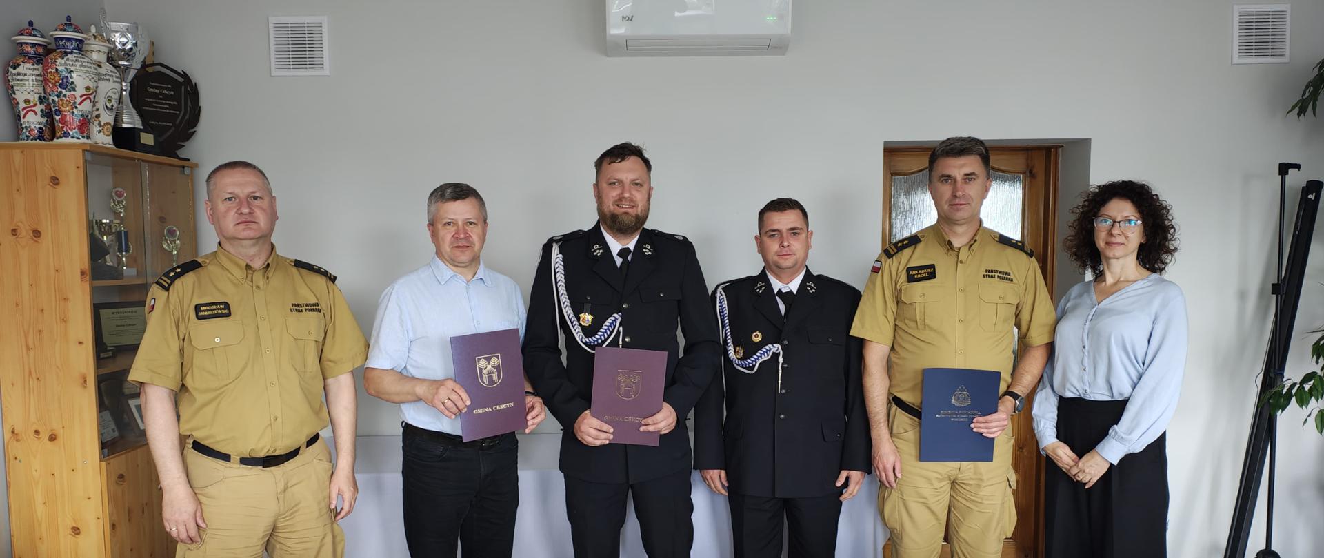 Sześć osób stoi w pomieszczeniu biurowym. Od lewej strony stoi strażak ubrany w mundur koloru piaskowego. Obok stoi mężczyzna ubrany w jasnoniebieską koszulę z krótkim rękawem i czarne spodnie. Mężczyzna trzyma w rękach brązową teczkę. Obok stoi dwóch strażaków ubranych w mundur wyjściowy koloru czarnego. Jeden z nich trzyma w rękach brązową teczkę. Obok stoi strażak ubrany w mundur koloru piaskowego. Strażak trzyma w jednej ręce niebieską teczkę. Obok strażaka stoi kobieta. Kobieta jest obrana w niebieską bluzkę i czarne spodnie.
Z przodu widać kawałek stołu i krzesła. Z lewej strony w rogu widać drewniany regał. Na regale stoją kolorowe wazony. Za szklanymi drzwiczkami regału widać puchary. Z prawej strony w tyle widać drzwi.
