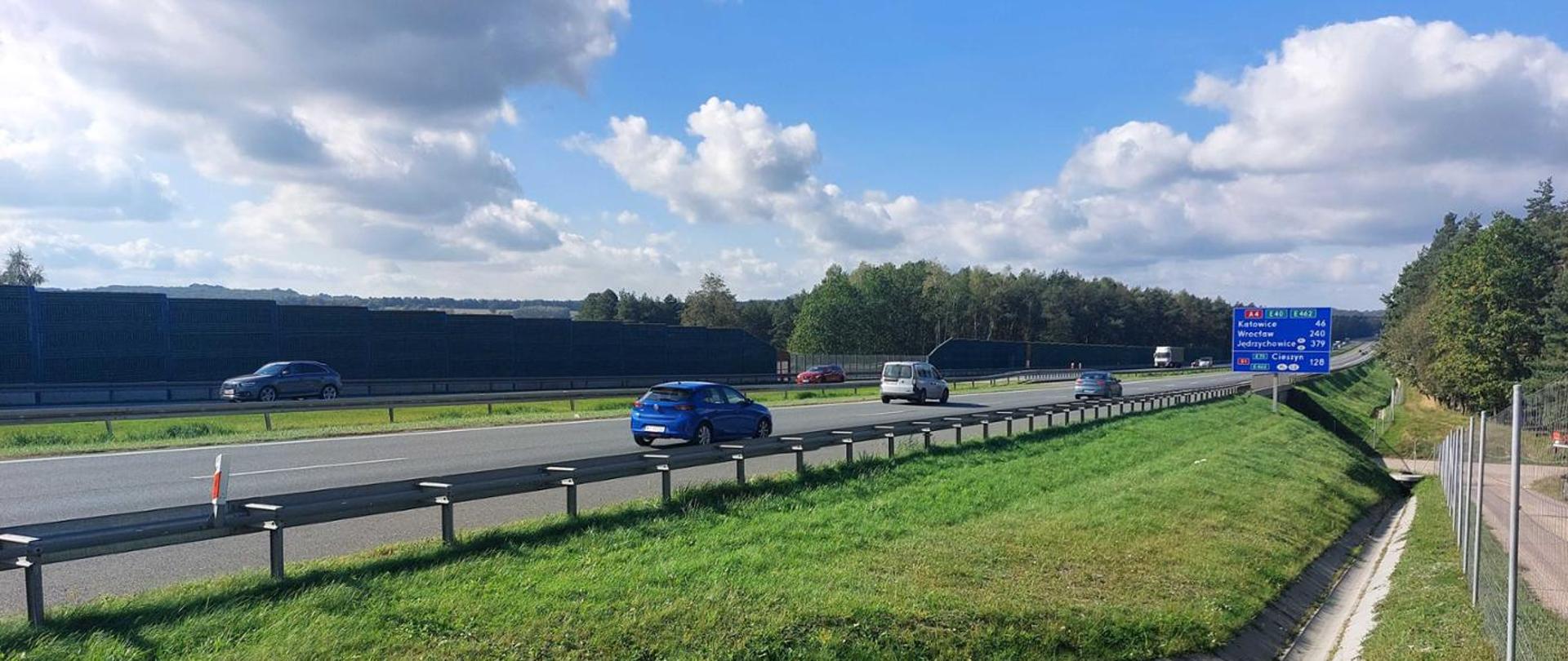 Dwupasmowa autostrada otoczona zielonymi pasami trawy; po obu kierunkach porusza się kilka samochodów. Po prawej stronie stoi niebieska tablica drogowa z kierunkami jazdy, a w tle widać las, ogrodzenie oraz fragmenty ekranów akustycznych. Na niebie znajdują się liczne białe, rozproszone chmury.