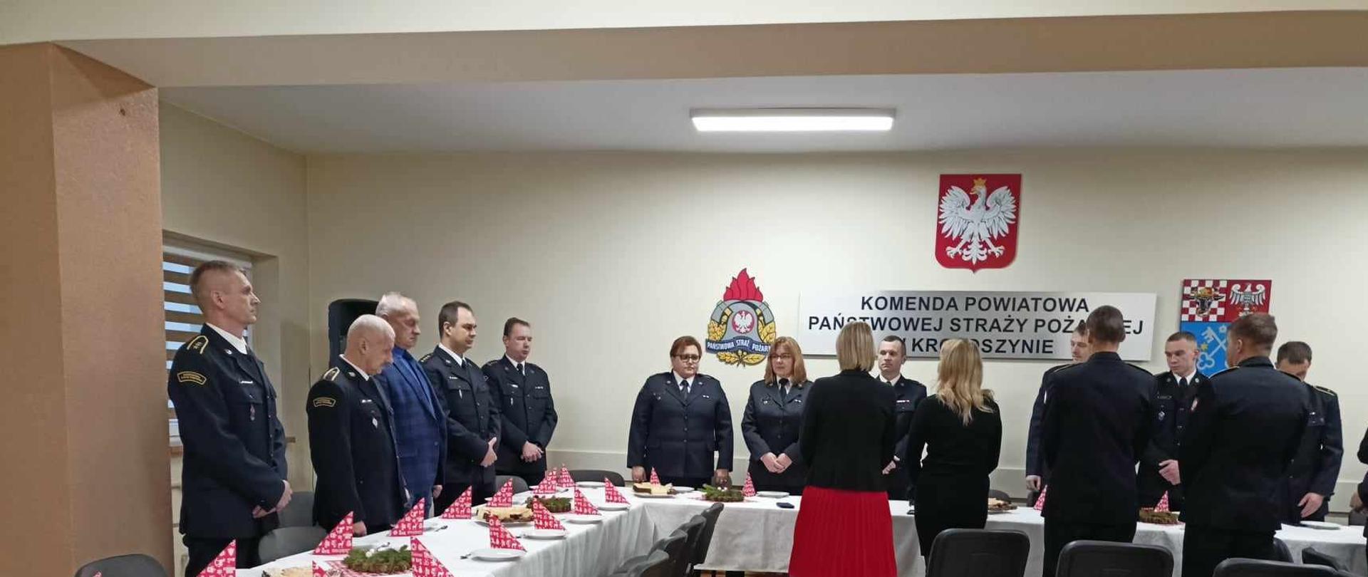 Na świetlicy krotoszyńskiej komendy zebrali się funkcjonariusze by w tym przedświątecznym czasie złożyć sobie życzenia. Stoły udekorowano białym obrusem, na których przygotowano drobny poczęstunek. Strażacy stoją przy stole wysłuchując okolicznościowych życzeń komendanta.