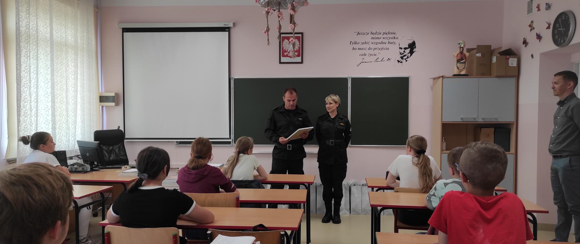 Zdjęcie przedstawia dwóch funkcjonariuszy Komendy Miejskiej Państwowej Straży Pożarnej w Rzeszowie podczas wręczania upominków za zajęcie II miejsca w konkursie Komendanta Głównego Państwowej Straży Pożarnej BEZPIECZNY JA uczniom klasy 7 w Szkoły Podstawowej w Kąkolówce. Strażacy stoją w klasie przed uczniami. Jeden z nich trzyma w ręce dyplom. 