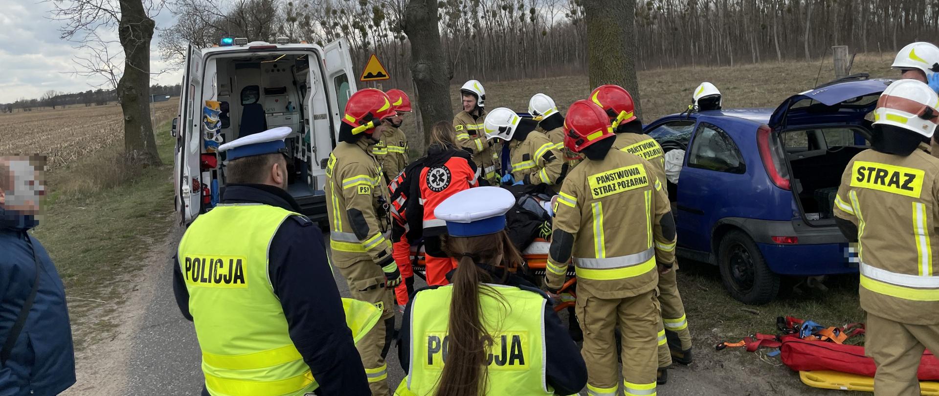 niebieski samochód z otwarta klapą bagażnika wokół niego strażacy w czerwonych i białych hełmach oraz funkcjonariusze Policji przed nimi karetka pogotowia wokół drzewo i jedna osoba postronna
