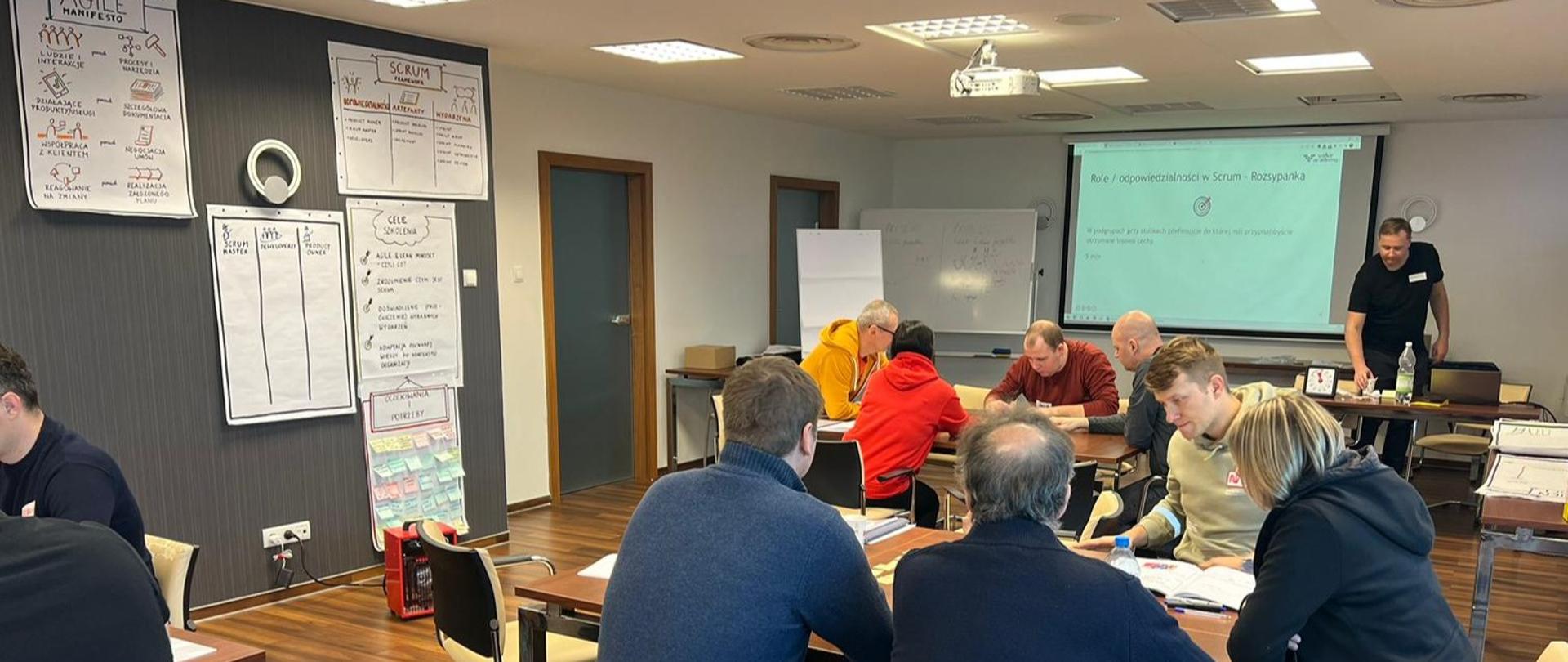 Na sali siedzą uczestnicy szkolenia w grupach, prowadzący stoi z przodu sali