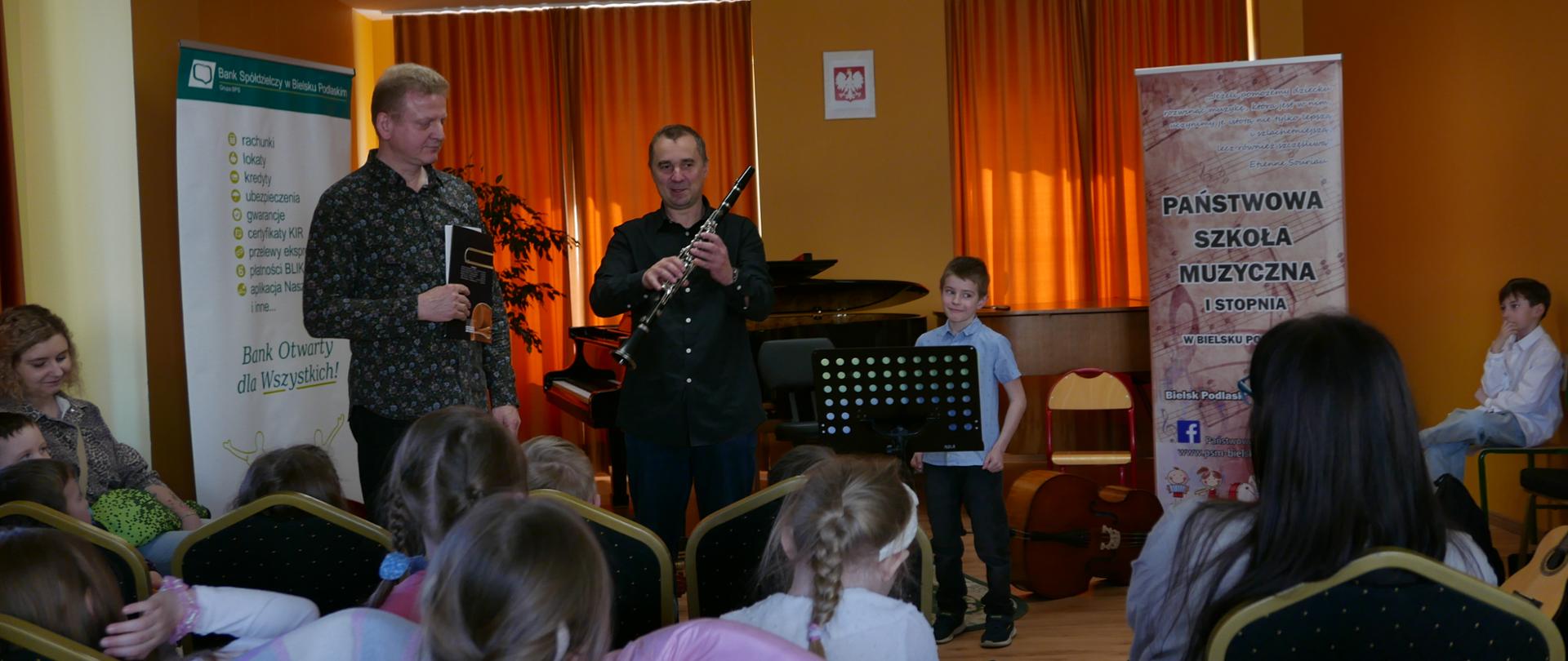 Widzimy publiczność złożoną z przedszkolaków Przed nimi dwóch panów prezentuje instrumenty muzyczne, jeden z nich trzyma klarnet i opowiada coś o nim. Obok stoi mały chłopak który za chwilę zagra przedszkolakom na klarnecie. 