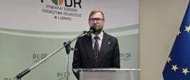 Mężczyzna w garniturze stoi przed mikrofonem na mównicy