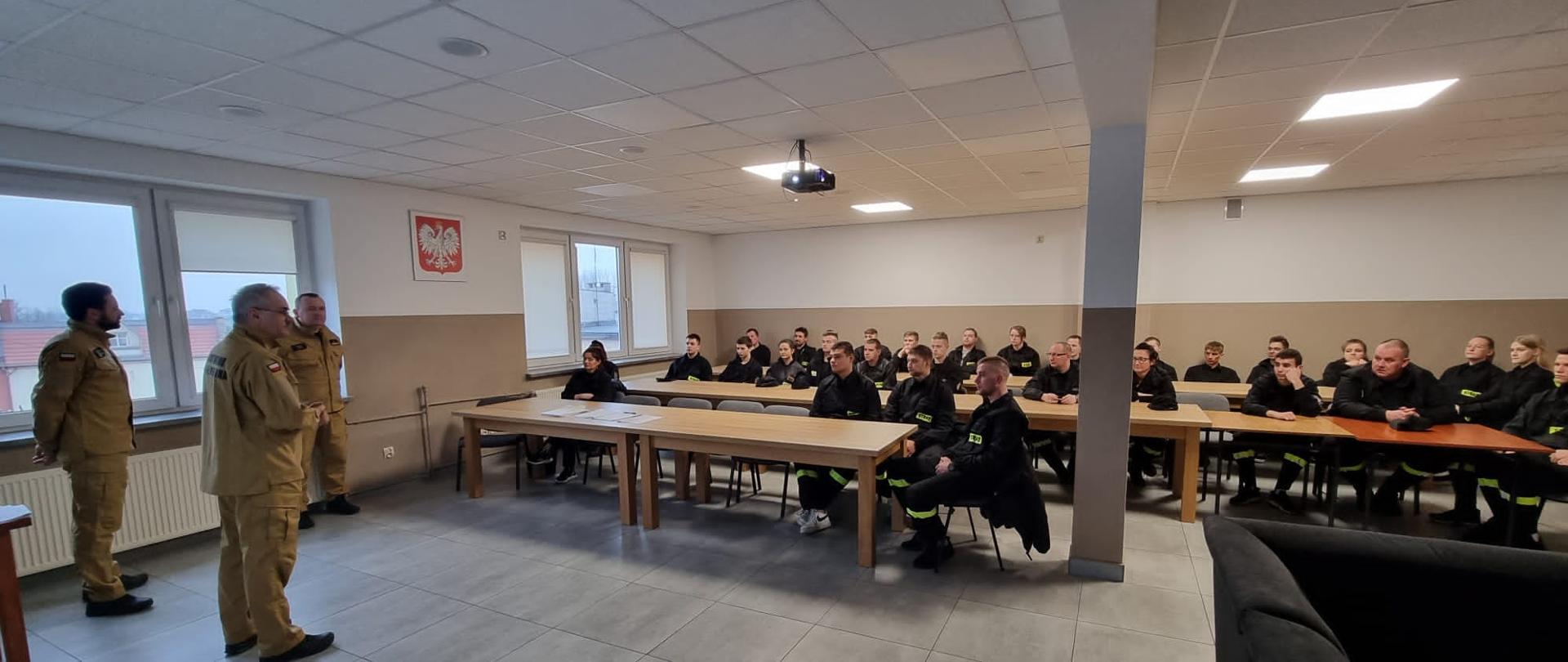 Kursanci siedzą na krzesłach. Przemawia Komendant Powiatowy PSP. Obok niego funkcjonariusze komendy