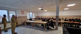 Kursanci siedzą na krzesłach. Przemawia Komendant Powiatowy PSP. Obok niego funkcjonariusze komendy