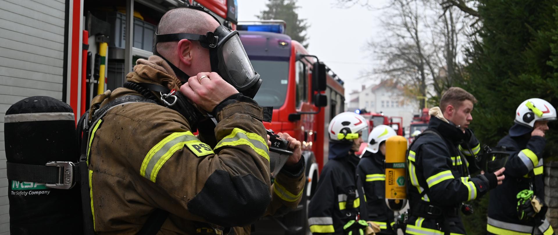 Po lewej stronie strażak ubiera maskę do aparatu powietrznego, ubrany jest w ubranie specjalne na plecach ma butlę z powietrzem, po prawej stronie stoi czterech strażaków, w mundurach specjalnych, jeden ma założony noszak z butlą w tle widać pojazdy pożarnicze.