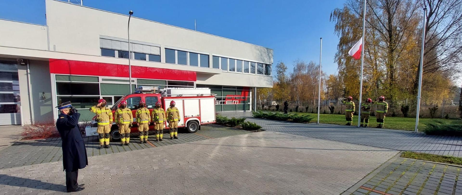 Na zdjęciu strażacy oraz komendant powiatowy PSP w Śremie w tle samochód ratowniczy JRG za nim budynek Komendy oraz Maszt na ktory strażacy wciągają Flagę Państwową 