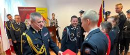 Pomorski komendant wojewódzki Państwowej Straży Pożarnej podaje czerwoną teczkę przedstawicielowi Młodzieżowej Drużyny Pożarniczej obok stoją zaproszeni goście.