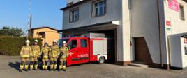 Strażacy stojący przed samochodem pożarniczym. Po prawej stronie zdjęcia budynek strażnicy. Po lewej stronie w tle budynek mieszkalny.
