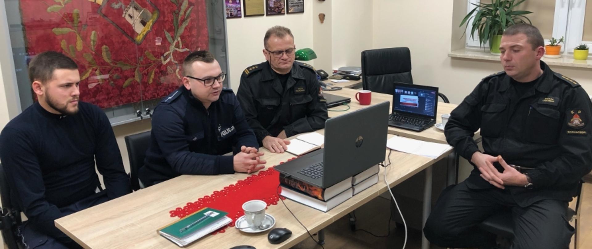 Na zdjęciu widocznych dwóch funkcjonariuszy policji (od lewej strony) w granatowym umundurowaniu oraz dwóch strażaków w czarnych ubraniach dowódczo-sztabowych. Trzech z nich siedzi przed laptopem i patrzą w ekran komputera.
