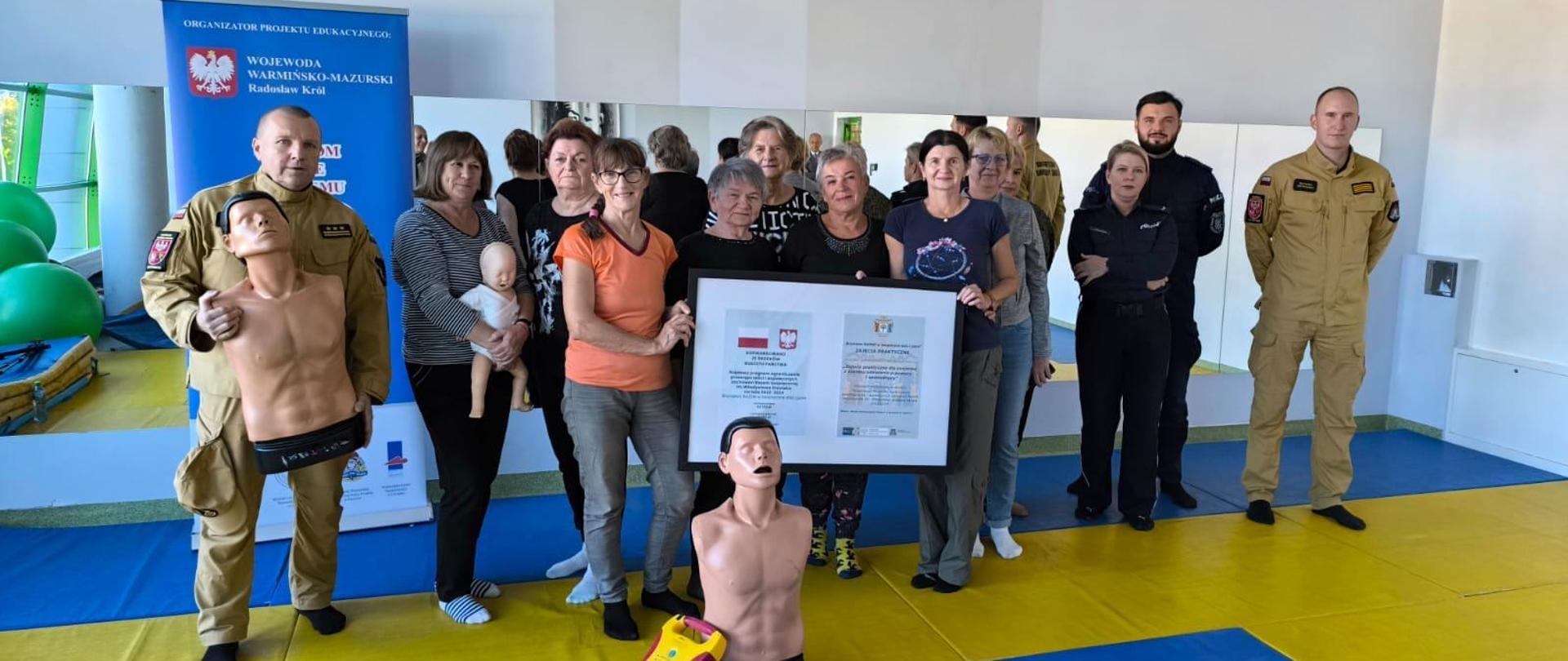 Na zdjęciu na sali sportowej funkcjonariusze szkolący i seniorki, uczestniczki szkolenia z pierwszej pomocy.