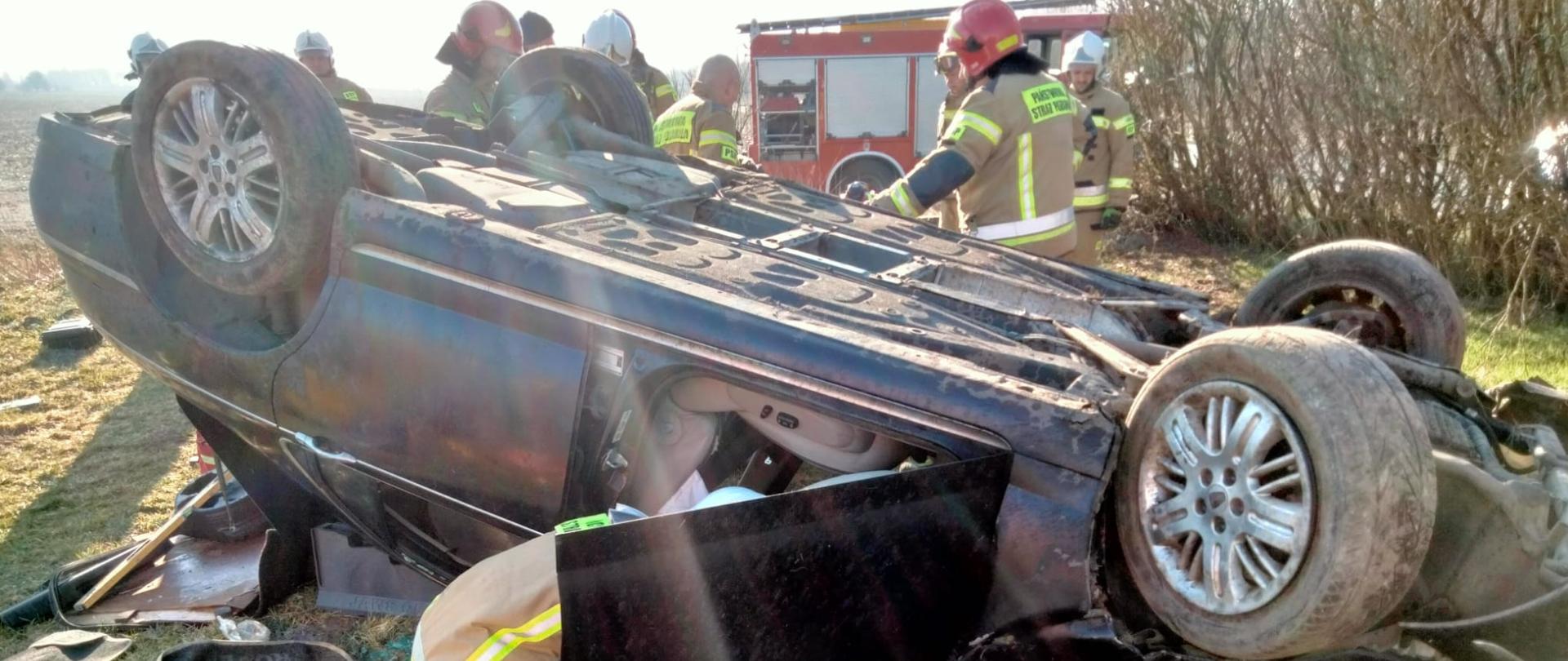 Fotografia przedstawia leżący na dachu samochód osobowy, w tle za wrakiem widoczny pojazd pożarniczy, przy pojeździe pracują strażacy w ubraniach specjalnych i w hełmach. Jest dzień, słonecznie.