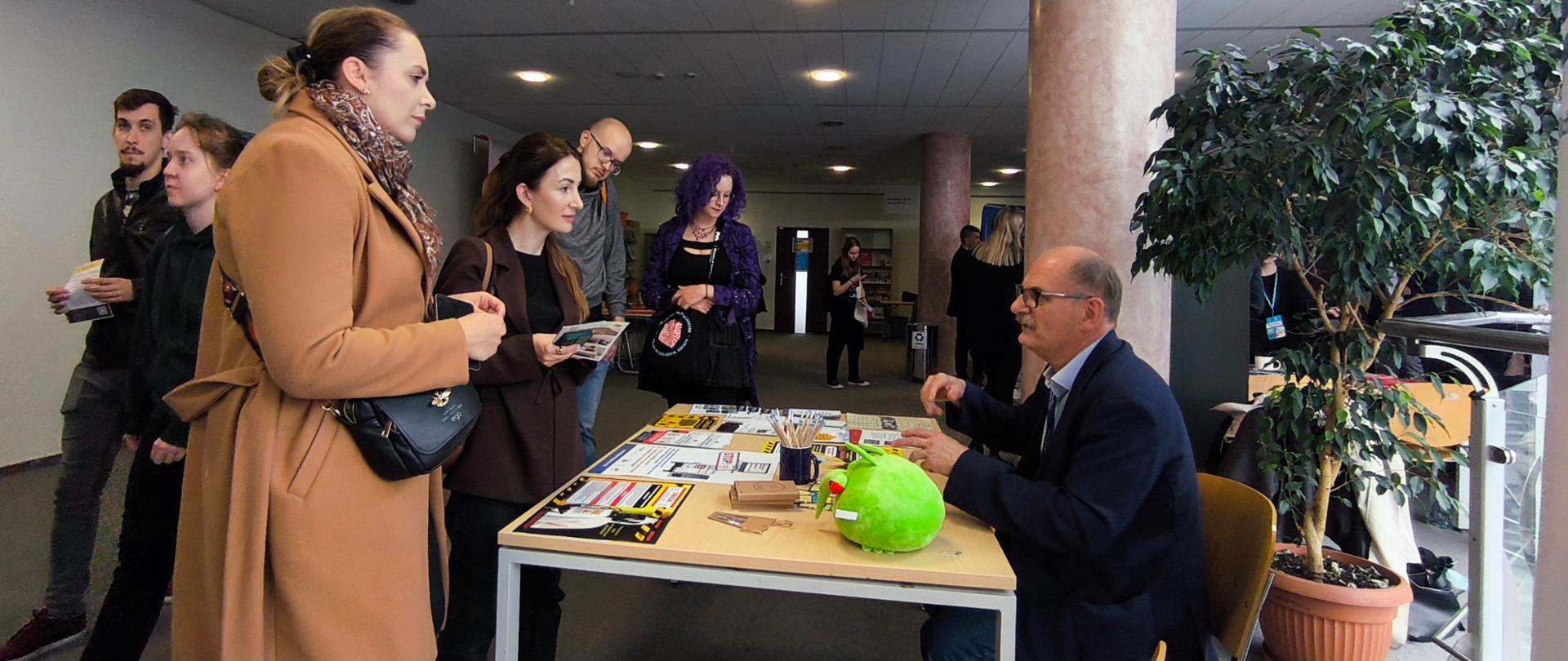 mężczyzna siedzący przy biurku wypełnionym gadżetami materiałami informacyjnymi rozmawia ze stojącą naprzeciw grupą młodych osób