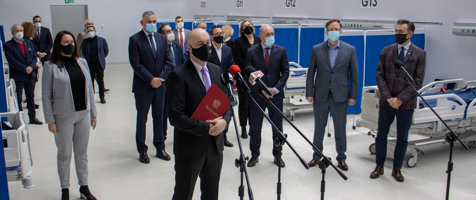 Konferencja prasowa Wojewody Opolskiego Adriana Czubaka. W centralnej części widać wojewodę przemawiające do publiczności, Przed nim cztery statywy, W tle osoby zaangażowane w powstanie szpitala.